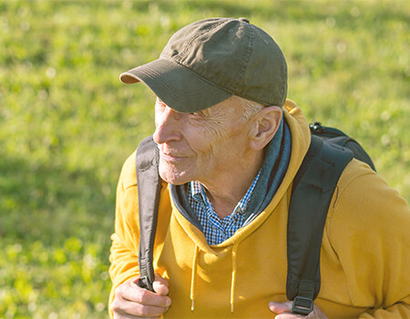 Senior man with backpack hiking outdoors in nature, wearing yellow hoodie and cap, embracing active aging